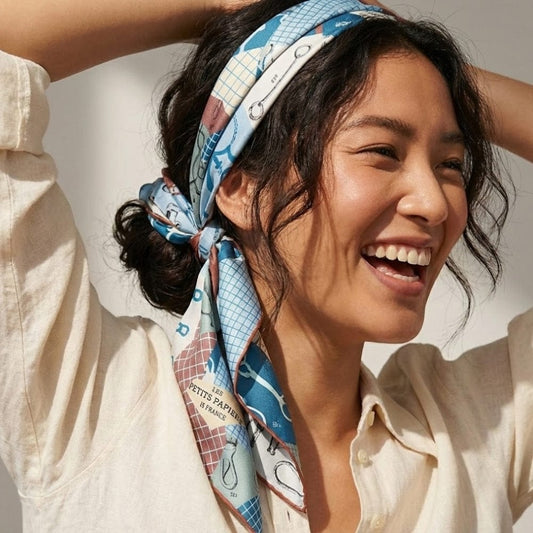 Foulard noué en satin bleu clair avec motifs graphiques porté par une femme souriant en blouse beige