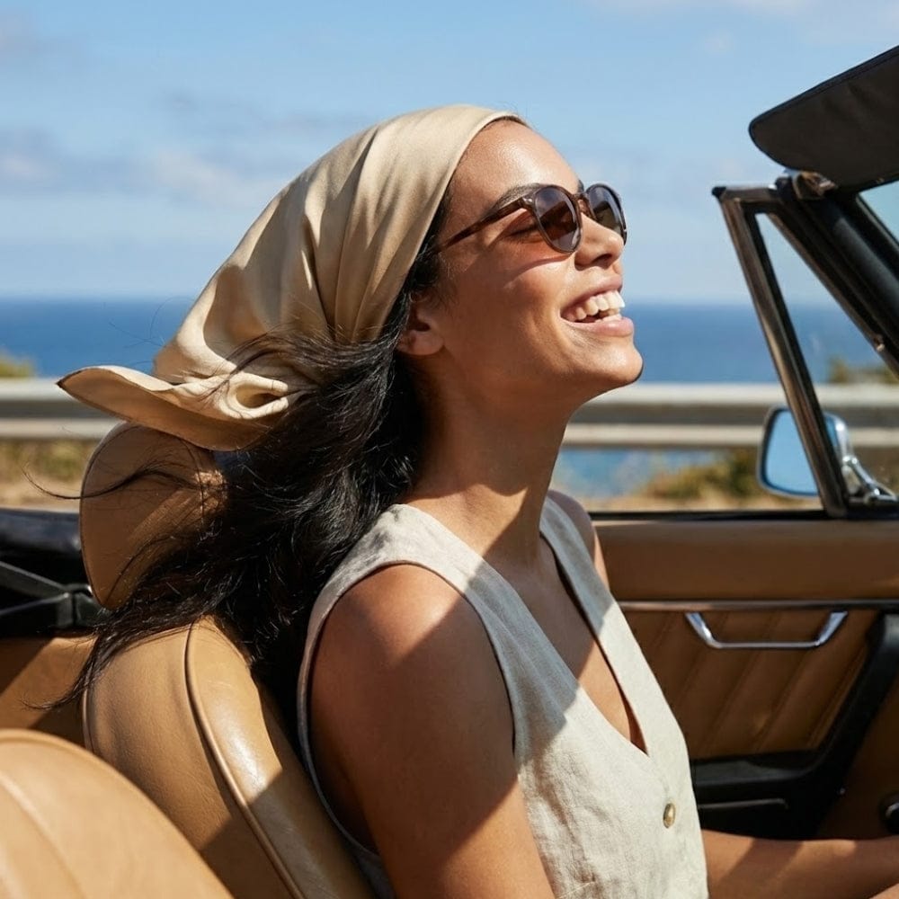 Foulard beige en satin noué sur la tête d'une femme souriante, assise dans une voiture décapotable au bord de la