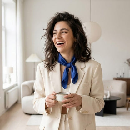 Foulard en soie bleu profond noué autour du cou d'une femme souriante, portant un blazer beige dans un intérieur moderne