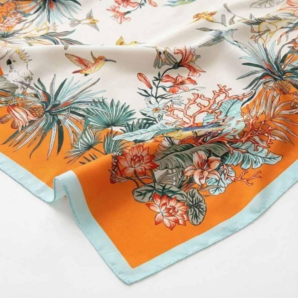Foulard carré en satin aux motifs floraux colorés sur fond crème et bordure orange, posé sur une surface blanche