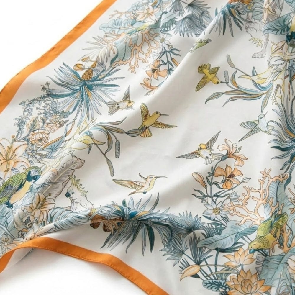 Foulard en soie blanc avec motifs floraux et oiseaux colorés, bord orange, posé sur une surface douce