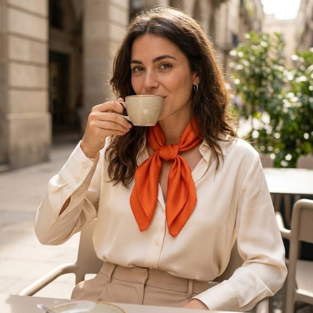 Foulard en soie orange noué élégamment autour du cou d'une femme, assise en terrasse avec une tasse de café