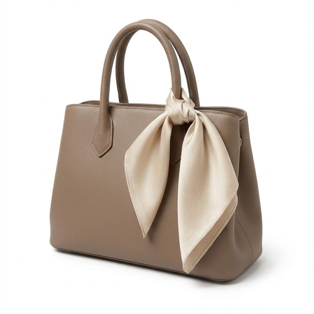 Foulard en satin beige noué sur un sac à main en cuir taupe, sur fond blanc